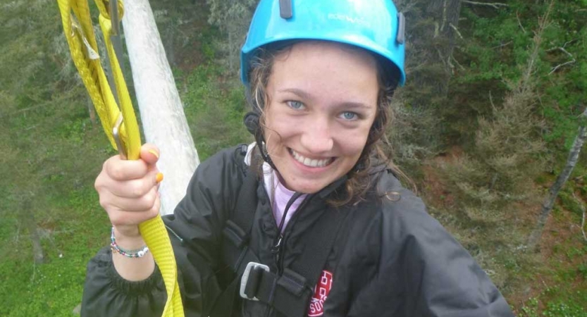 a student wearing a helmet smiles at the camera while participating in a high ropes course with outward bound